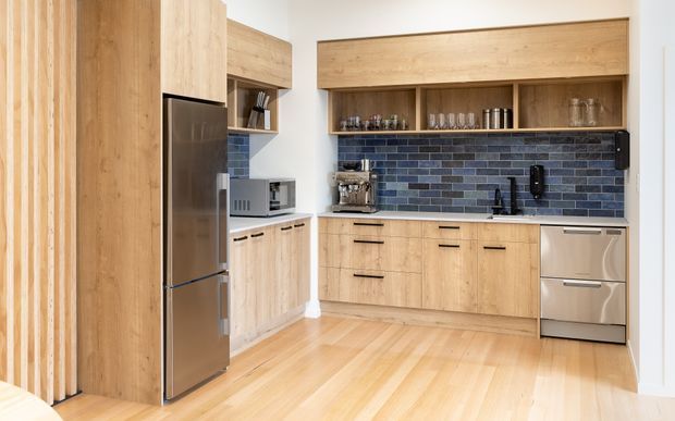 Bright contemporary kitchen featuring extensive light timber cabinetry, integrated open shelving, a dark blue-grey brick-pattern tiled splashback, stainless-steel refrigerator and dishwasher, and pale timber flooring. Vertical timber slats and a compact appliance nook add architectural detail.