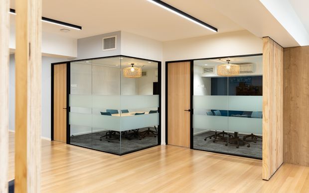 A bright contemporary office interior with two enclosed glass meeting rooms featuring black-framed glazing, frosted privacy bands, timber doors, and visible conference tables and office chairs. The space has light timber flooring, pale walls and ceilings, linear recessed lighting, and vertical timber slatted detailing around the openings.