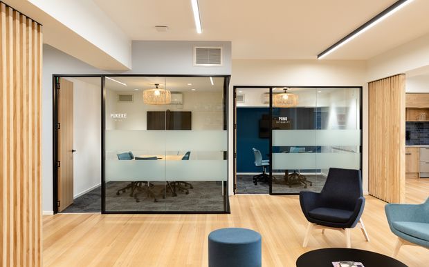 Contemporary office reception or breakout area with pale timber flooring, black-framed glazed meeting rooms, frosted privacy bands, timber slatted screens, and modern lounge seating. A kitchenette is partially visible at right, while illuminated linear ceiling lights and pendant fixtures provide a clean professional fit-out.