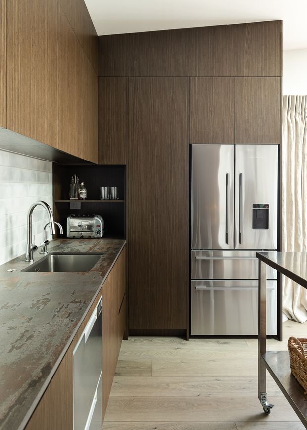A compact contemporary kitchen with tall dark timber-grain cabinetry, integrated storage, a stainless-steel refrigerator, and a stone-look countertop with an undermount sink and brushed metal tapware. A white tiled splashback, open shelf with small appliances, pale timber flooring, and a narrow timber-framed island or table complete the space. Natural light enters through curtains at the right.