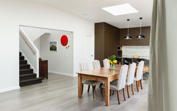 Bright open-plan dining and kitchen interior with a large timber dining table surrounded by upholstered chairs. The space features pale timber-look flooring, white walls, a dark timber kitchen with a light stone splashback, black pendant lights, a skylight, and an adjacent carpeted staircase. Contemporary furnishings include colourful wall art and a floral table arrangement.