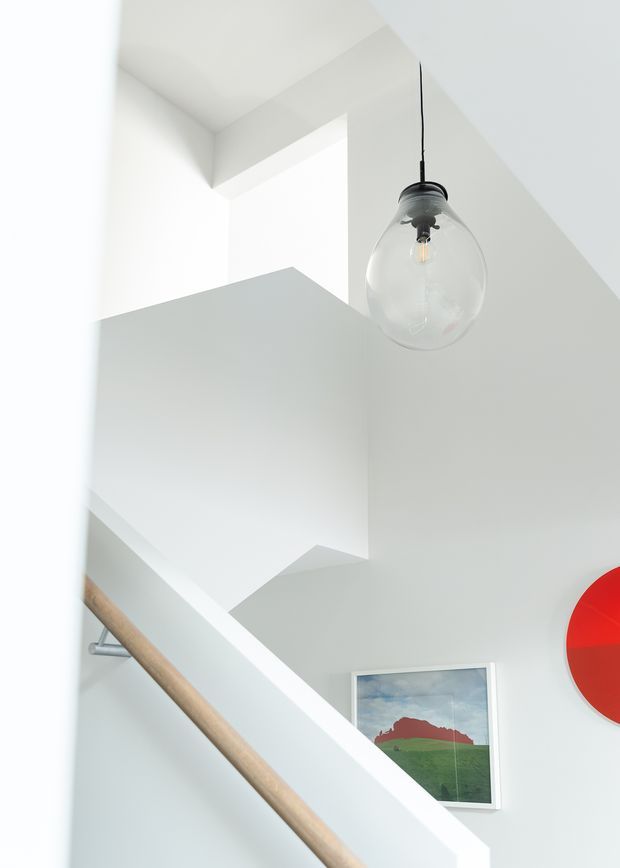 Bright minimalist stairwell with crisp white walls and angular geometric ceiling and stair forms. A light timber handrail with metal supports runs diagonally through the lower portion, while a clear glass pendant light hangs from the high ceiling. Framed artwork and a bold red circular wall element add small accents.