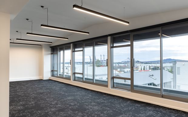 A contemporary, empty commercial office interior with dark patterned carpet, white walls and ceiling, suspended linear pendant lights, and an extensive wall of dark-framed windows overlooking an urban waterfront or harbour setting. The space is bright, open, and minimally finished.