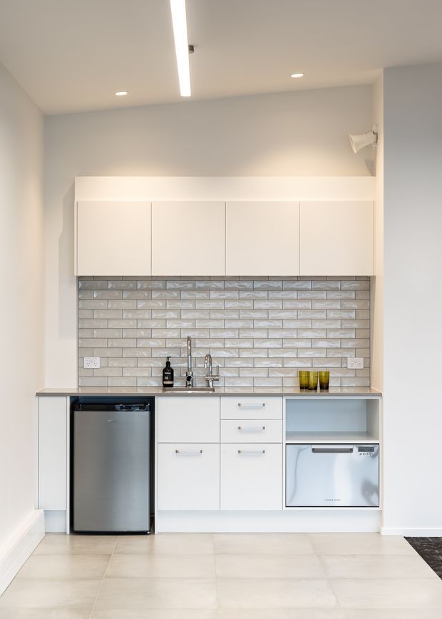 A compact, clean-lined kitchenette with white flat-panel cabinetry, a grey engineered-looking benchtop, glossy light grey subway-tile splashback, stainless-steel under-bench refrigerator and dishwasher, and a small stainless-steel sink. Recessed ceiling lights and a linear LED strip provide bright, even illumination. The pale tiled floor and neutral finishes create a minimalist, contemporary appearance.