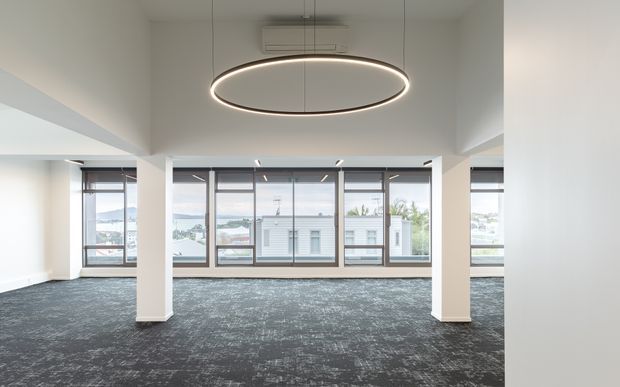 Open-plan commercial interior with a broad carpeted floor, white structural columns and walls, extensive floor-to-ceiling glazing, and a large circular pendant light. The windows overlook neighbouring buildings and a waterfront or harbour-like setting. The space appears vacant and suitable for office use.