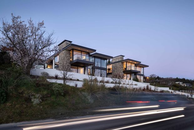 Twilight exterior photograph of a large contemporary multi-level residence on a landscaped sloping site. The building has strong horizontal flat roof planes, extensive floor-to-ceiling glazing, projecting balconies, dark-framed windows, and prominent stone-clad vertical volumes. Long-exposure vehicle light trails run along the road in the foreground.
