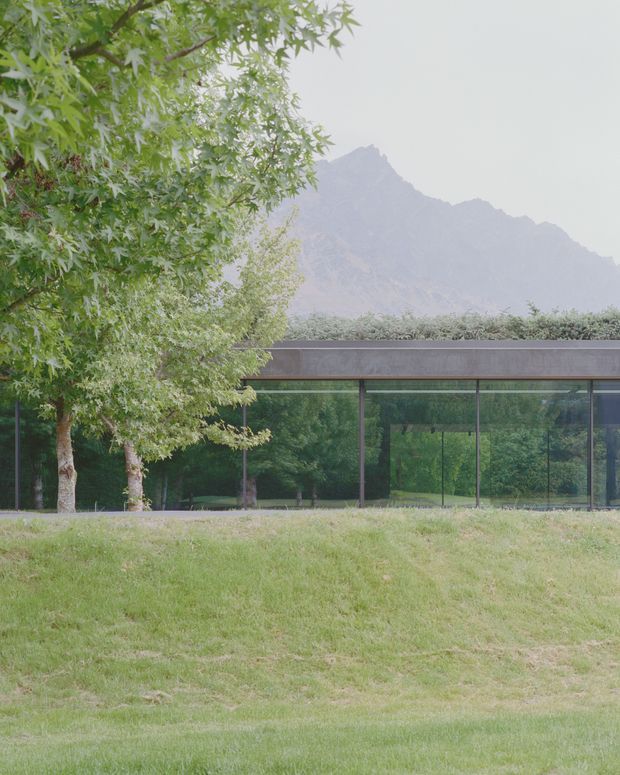 A low, elongated modern pavilion set within a landscaped grassy site. The building has extensive floor-to-ceiling glazing framed in dark metal, a thin horizontal roofline, and a transparent connection to the surrounding trees. Mature deciduous trees dominate the foreground, with a hazy rugged mountain ridge visible behind the structure. The image has a quiet, minimal, nature-integrated character.