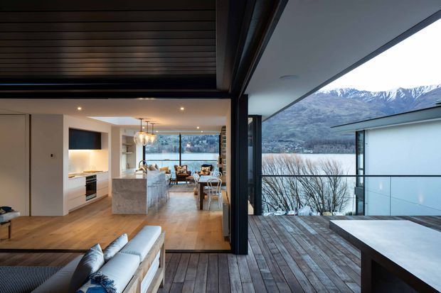 Open-plan contemporary home interior opening onto a large timber deck, with a kitchen and dining area visible beyond the living room. Floor-to-ceiling glazing and an expansive covered opening frame a mountainous landscape. The space features warm timber flooring, dark timber ceiling slats, white cabinetry, a stone kitchen island, and a neutral upholstered sofa.