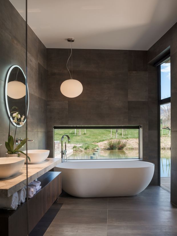 A contemporary bathroom with dark large-format wall and floor tiles, a freestanding white oval bathtub positioned beneath a wide horizontal window, and a long double vanity with white vessel basins and timber cabinetry. A round illuminated mirror, suspended globe pendant, black-framed glazing, and views to a pond and landscaped grounds complete the calm, refined space.