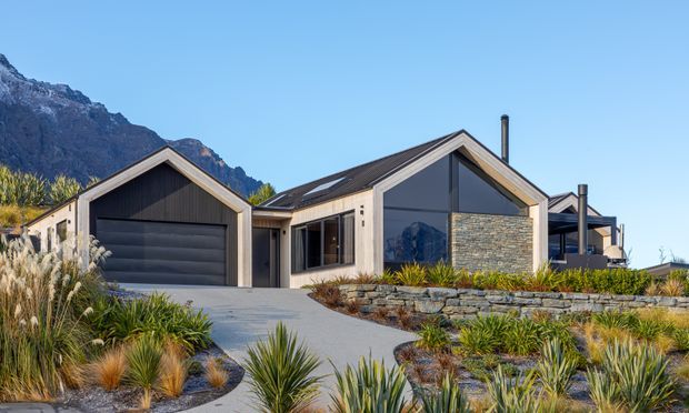 Contemporary single-storey gabled home with dark metal roofing, light rendered and stone-clad walls, large glazed gable ends, and an integrated garage. The house is set against a dramatic mountain backdrop and surrounded by low-maintenance native-style landscaping, stone retaining walls, and a concrete driveway.