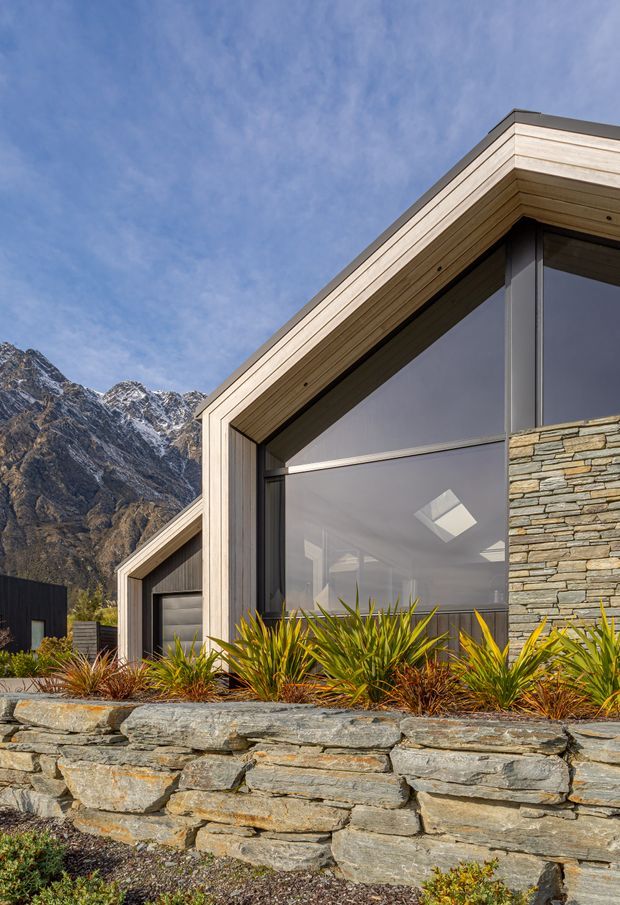 Exterior architectural photograph of a contemporary mountain home with steeply angled roof forms, expansive dark-framed glazing, pale timber-lined fascias, and layered natural stone walls. Spiky landscaped planting fronts the building, with snow-dusted rugged mountains visible behind it.
