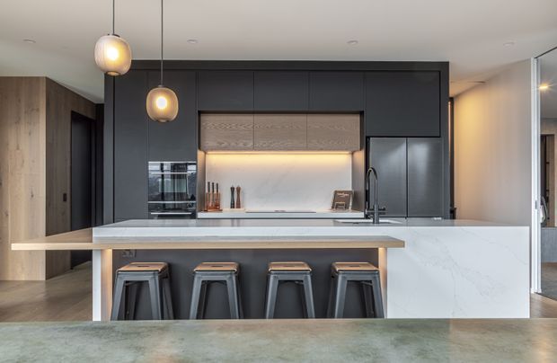 A contemporary kitchen with a large island and four black bar stools. The space features dark matte cabinetry, pale timber-look overhead panels, a white veined stone benchtop and splashback, integrated stainless-steel appliances, warm pendant lighting, and timber flooring.