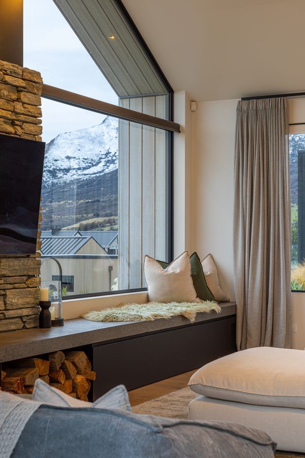 A warm, refined living room with a large corner window framing a snow-covered mountain landscape. The room features a substantial stacked-stone fireplace with integrated firewood storage, a dark built-in window seat with cushions, floor-length neutral curtains, and layered upholstered seating in the foreground. The architecture combines generous glazing with natural stone and timber-toned elements.