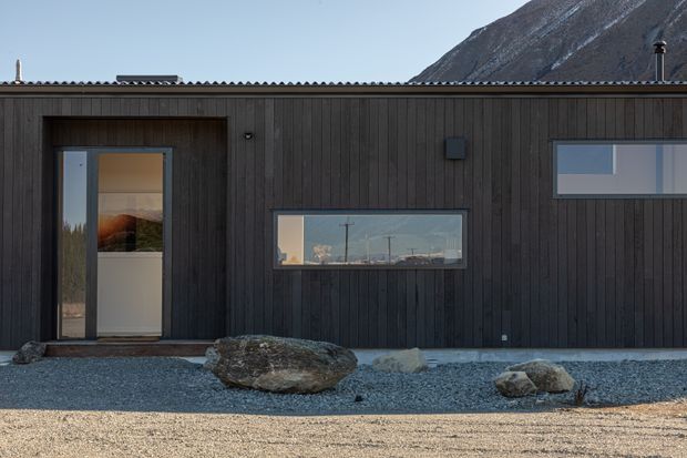 A minimalist single-storey building facade clad in dark vertical timber boards, with a low corrugated roof, narrow horizontal windows, and a glazed entry door. The building sits in a gravel landscape with large natural rocks, backed by a rugged mountain slope. The dark facade contrasts with the pale gravel and clear sky.