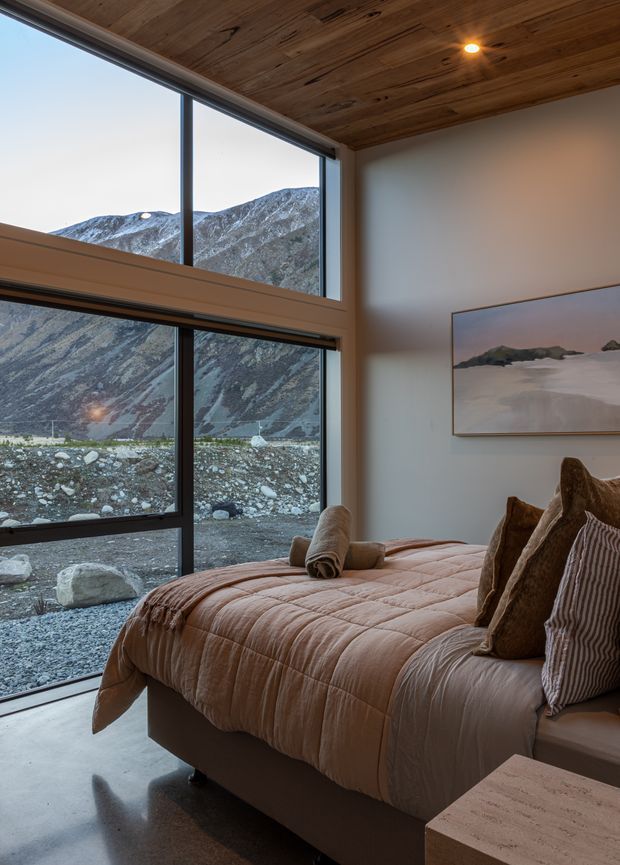 A contemporary bedroom with a low upholstered bed dressed in warm taupe and brown bedding, positioned beside expansive black-framed floor-to-ceiling glazing. The room features a warm timber-lined ceiling with recessed lighting, a smooth neutral wall, polished grey concrete flooring, and a minimalist landscape artwork. Snow-dusted rugged mountains and a rocky landscape are visible through the windows.