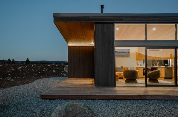 Exterior dusk view of a compact contemporary single-storey pavilion with dark vertical timber cladding, a projecting flat roof, warm timber soffit, and extensive floor-to-ceiling glazing. A timber deck fronts the building, while the illuminated interior reveals a living area and kitchen. The setting appears sparse and rocky, with a restrained indoor-outdoor connection and warm evening lighting.