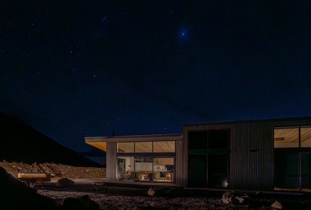 A low, single-storey modern residence photographed at night beneath a star-filled sky. The house has a strongly horizontal composition, dark vertically ribbed exterior cladding, extensive floor-to-ceiling glazing, and warm interior lighting visible through the windows. A dramatic mountain silhouette and rugged rocky terrain surround the building.