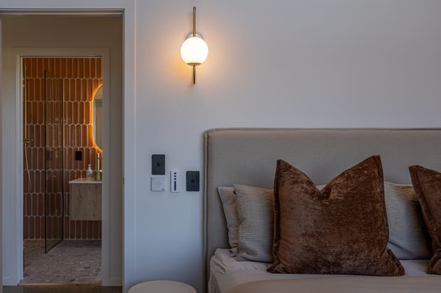 A warmly lit bedroom interior with a large upholstered bed and layered neutral and brown pillows. A minimalist wall sconce is mounted above the bed, while an open doorway reveals a bathroom or dressing area with distinctive vertical terracotta-toned tilework and a stone-like floor. The room has a refined, contemporary character with soft grey walls and tailored detailing.