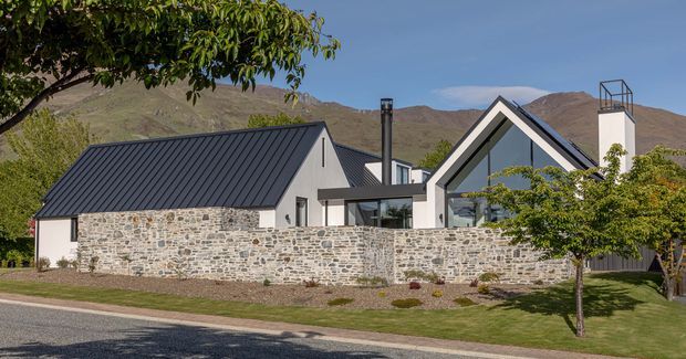 Exterior view of a contemporary gabled house in a mountainous setting. The home combines pale rendered walls, dark standing-seam metal pitched roofs, extensive natural stone boundary walls, large glazed gable sections, and a prominent chimney. Mature landscaping and a lawn sit in front of the property, with rugged hills visible behind.
