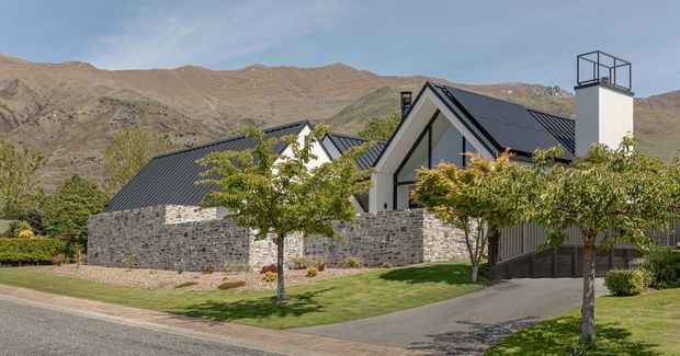 Exterior view of a contemporary alpine residence set against a rugged, dry mountain landscape. The home has steep gabled roof forms clad in dark metal, pale rendered gable walls, extensive grey natural-stone masonry walls, a tall chimney with a metal cap, and landscaped lawns with young deciduous trees. A paved driveway and street edge are visible in the foreground.