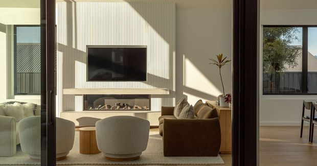 A sunlit contemporary living room viewed through large glazed openings, with a sculptural white ribbed feature wall incorporating a wall-mounted television and linear fireplace. The room contains rounded boucle armchairs, a brown sofa, a low timber coffee table, and a neutral rug. Strong diagonal sunlight and shadows create a warm, refined atmosphere. A dining area and exterior garden are visible beyond the living room.