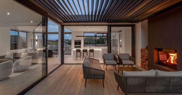 A refined indoor-outdoor living area with extensive floor-to-ceiling glazing, timber flooring, a dramatic slatted timber ceiling, contemporary lounge furniture, and a built-in fireplace with stacked firewood. The space opens visually toward a bright kitchen and dining area beyond, with warm evening lighting and views of the surrounding landscape.