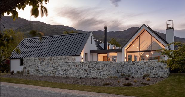 Exterior view of a contemporary alpine residence at dusk, featuring steep gabled roof forms, dark standing-seam metal roofing, pale plaster walls, extensive warm-lit glazing, and a low natural stone boundary wall. Dramatic mountain terrain forms the backdrop, with landscaped grounds and a paved street in the foreground.