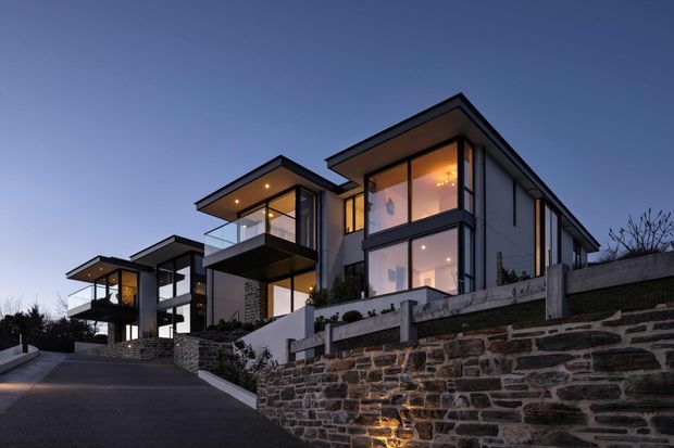 Twilight exterior view of a contemporary multi-storey residential development on a sloped site. The homes feature angular flat roof forms, extensive floor-to-ceiling glazing, projecting upper-level balconies, dark-framed windows, and pale rendered or clad facades. A substantial stepped natural-stone retaining wall and landscaped terraces define the foreground.