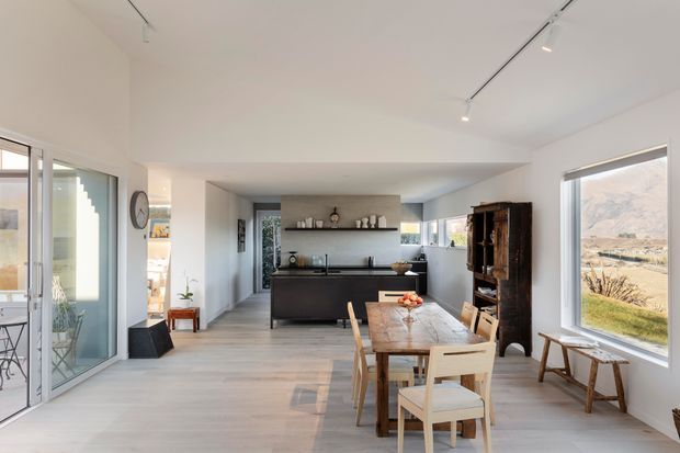 Bright open-plan interior combining a kitchen, dining area and living space. The room has white walls and ceiling, pale timber flooring, a dark kitchen island with integrated cabinetry, a long dining table with timber chairs, and large glazed doors and windows overlooking the landscape. Track lighting and simple contemporary furnishings create an airy, calm atmosphere.
