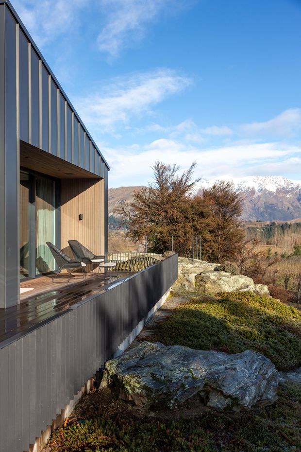 Exterior view of a contemporary, elongated mountain dwelling with dark vertical metal cladding and extensive floor-to-ceiling glazing. A timber-lined covered terrace with outdoor lounge chairs projects over a rugged landscaped site with exposed rocks and native vegetation, overlooking a dramatic snow-capped mountain range under a bright blue sky.