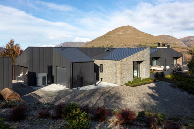 Exterior view of a contemporary, low-profile mountain residence composed of dark metal-clad gabled volumes and a central pale stone-clad section. The home sits within a gravelled landscaped site, with large windows, a sheltered outdoor terrace, and rugged hills visible behind it.
