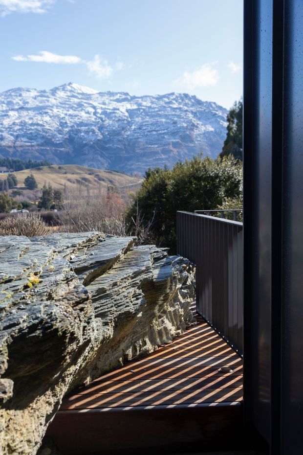 A close architectural view from beside a dark-framed opening onto a narrow timber deck or balcony. A rugged natural rock outcrop forms the edge of the deck, with dark vertical metal or timber balustrading. Beyond, a dry rolling landscape and snow-covered mountain range are visible under a clear blue sky.