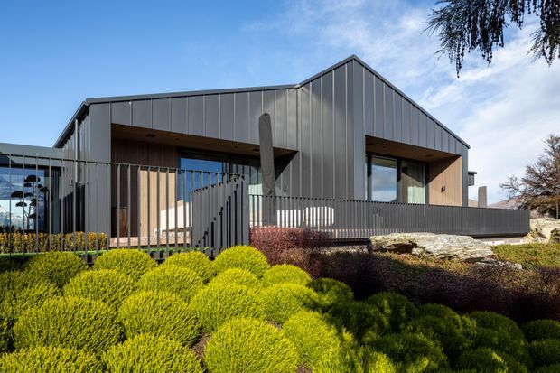 Contemporary single-storey residence with a sharply pitched gable roof and dark vertical metal cladding. Large recessed glazed openings and warm timber-lined soffits create sheltered outdoor areas, enclosed by a dark metal picket fence. The house sits within a carefully landscaped garden with sculpted shrubs and stone retaining elements under a clear blue sky.