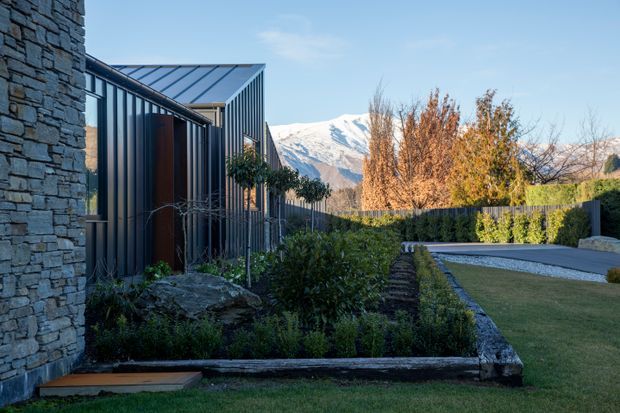 Exterior view of a contemporary dwelling and landscaped garden, with dark vertical metal cladding, a prominent rough stacked-stone wall, structured planting beds, lawn, and a gravel driveway. Snow-covered mountains are visible in the background.