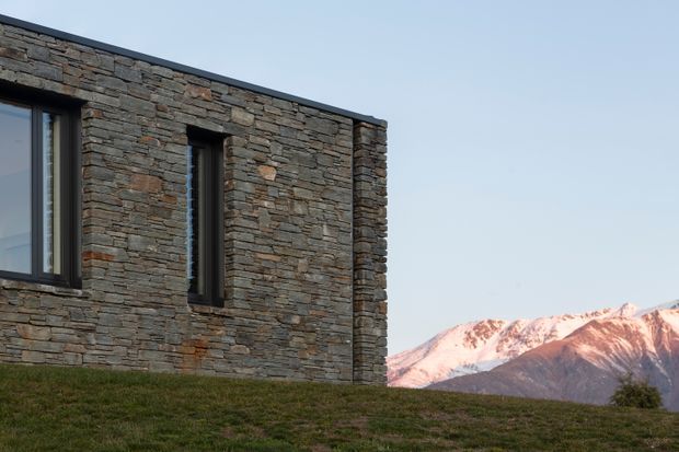 Exterior detail of a contemporary, low-profile building clad in irregular stacked natural stone, with narrow dark-framed windows and a simple flat roofline. The structure sits in a grassy alpine setting with snow-capped mountains visible in the background.