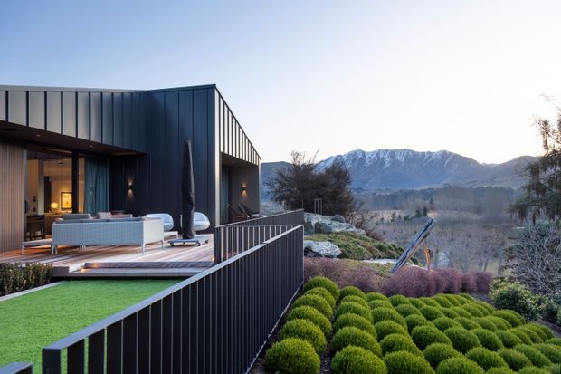 Contemporary mountain-view house exterior with a dark vertically ribbed metal-clad upper volume, expansive glazing, and a timber-toned outdoor living terrace furnished with a sofa and chairs. A dark metal balustrade frames a landscaped garden of clipped shrubs, with snow-dusted mountains visible in the distance.