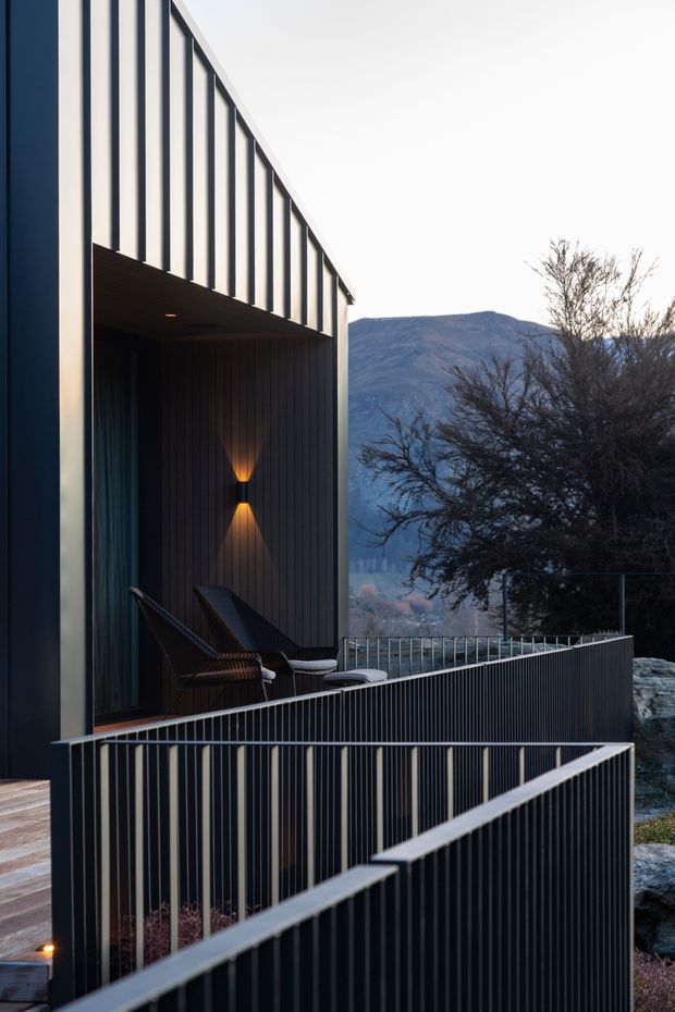 A contemporary multi-level exterior terrace or balcony attached to a dark-clad building, with strong vertical metal balustrades and an overhead projecting structure. Outdoor lounge chairs sit beneath a warm wall light, while a mountain landscape and trees are visible beyond the property. The composition emphasizes geometric lines, dark materials, and a dramatic dusk atmosphere.