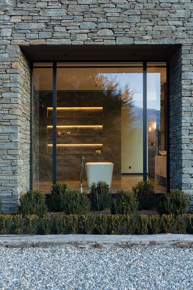 Exterior view of a contemporary stone-clad building with a deeply recessed, full-height glazed opening. Through the dark metal-framed glazing is a refined bathroom featuring a freestanding white bathtub, illuminated recessed wall shelving, dark stone wall finishes, timber flooring, and timber cabinetry. Low clipped shrubs and a gravel forecourt occupy the foreground, while reflections reveal a mountainous landscape beyond.