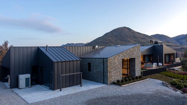 Contemporary single-storey residential building with multiple dark metal-clad volumes and pitched roof forms, connected by a central stone-clad section. The house sits in a gravelled landscape with low planting and is framed by rugged mountains in the background. Large glazed openings and warm interior lighting are visible on the right-hand facade.