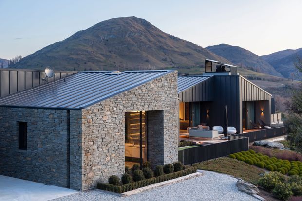 Exterior view of a contemporary mountain house composed of rough stacked-stone walls, dark vertical metal cladding, and low-pitched metal roofs. Large glazed openings reveal warm interior lighting and seating, while formal landscaping and a pale gravel forecourt frame the building. A rugged mountain landscape rises directly behind the home.
