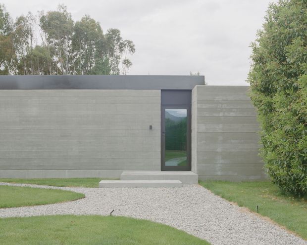 A minimalist single-storey concrete building exterior with broad horizontal board-formed concrete walls, a dark flat roof edge, and a narrow glazed entry door. A light gravel path leads through manicured lawn toward the entrance, with mature trees and hedging surrounding the structure.