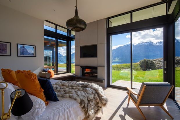 A bright, contemporary living room with floor-to-ceiling black-framed glazing overlooking a dramatic mountain and lake landscape. The room features a built-in fireplace and television wall, pale timber flooring, a large white sofa layered with orange and navy cushions and a sheepskin throw, a timber-framed lounge chair, and a sculptural pendant light. High-level internal glazing and strong indoor-outdoor visual connection add to the spacious, refined alpine character.