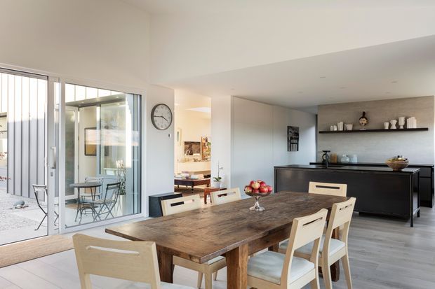 Open-plan contemporary dining and kitchen area with a large rustic timber dining table and light timber chairs in the foreground. The kitchen features dark cabinetry, a dark benchtop, a pale textured splashback or wall surface, and a long open display shelf. Full-height sliding glass doors connect the dining space to a small paved outdoor courtyard or patio, while a living area is visible beyond the dining zone.