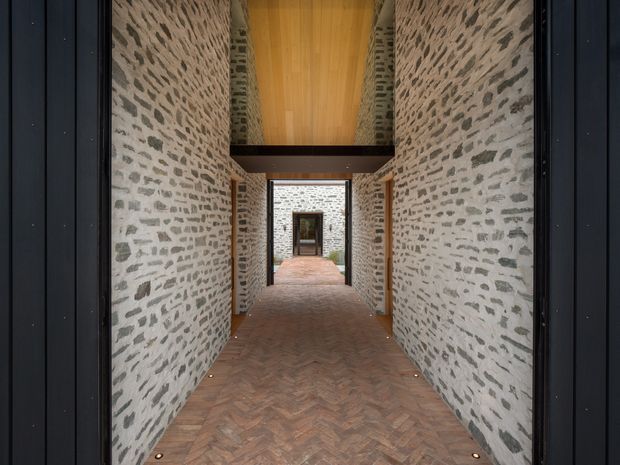 A long, narrow architectural passageway framed by dark metal openings, with exposed pale stone masonry walls on both sides, a warm timber-lined ceiling, dark structural beams, and a herringbone timber floor. The corridor leads through a series of aligned openings toward another stone courtyard-like space, with discreet floor-level lighting along the edges.