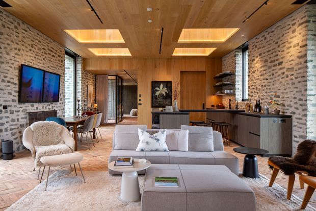 Open-plan contemporary living and kitchen interior with exposed light stone masonry walls, warm timber ceiling and ceiling beams, integrated recessed lighting, and herringbone timber flooring. The room includes a large light-grey sectional sofa, sculptural lounge chairs, a dining area, and a dark kitchen with island seating. Tall glazed openings and a layered, warm material palette create a refined yet inviting atmosphere.