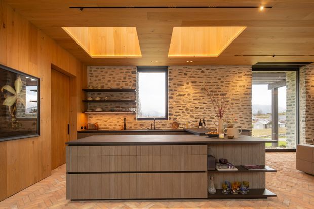 Warm contemporary kitchen with a large central island, integrated open shelving, timber-lined walls and ceiling, exposed rustic brick feature walls, recessed illuminated ceiling panels, and expansive windows overlooking a residential landscape. The island has textured wood cabinetry and a dark stone-like benchtop, with terracotta-toned flooring and understated styling.