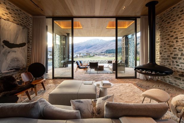 Warm, sophisticated living room with exposed rough stone walls, a timber-lined ceiling, and large black-framed glazed doors opening to a terrace with dramatic mountain views. The room features a sculptural suspended wood-burning fireplace, low contemporary sofas, a large upholstered ottoman, mid-century-inspired lounge chairs, and layered neutral textiles.