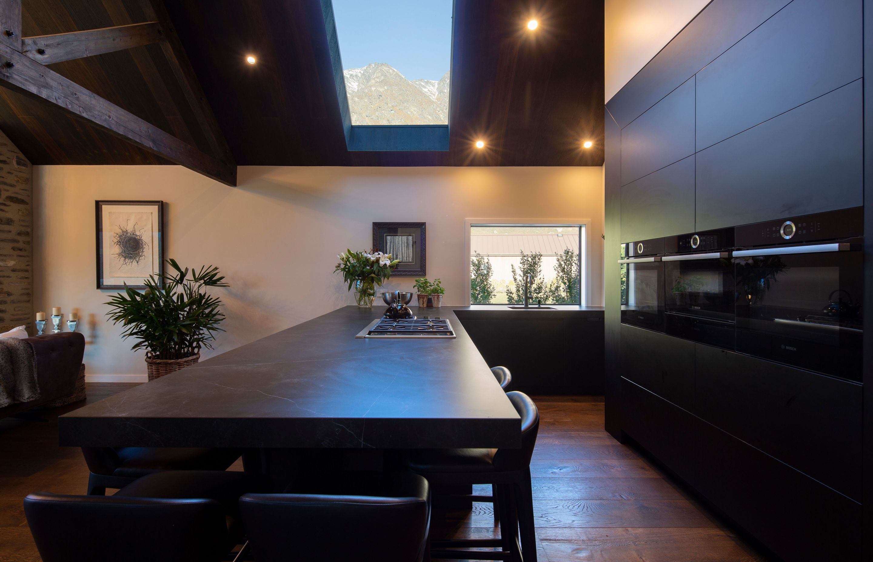 The kitchen features charcoal-toned cabinetry and a matte-black peninsula with a hob inset for meal preparation while chatting with friends. Through the skylight, the cook gets a peek at The Remarkables.