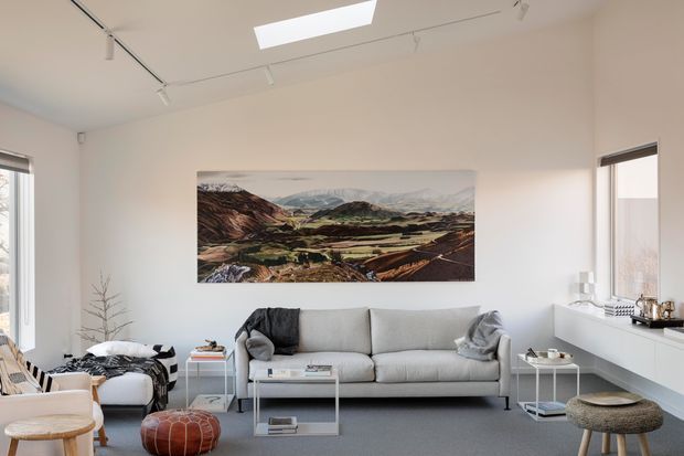 Bright, minimalist living room with a large light-grey sofa, white walls, grey carpet, low white coffee table, side tables, and a long white built-in window-side cabinet. Track lighting and a rectangular skylight provide illumination, while a large landscape artwork forms the main wall feature. A lounge chair and ottomans add seating, with windows on both sides.