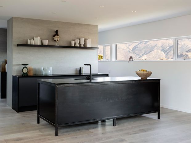 A bright contemporary kitchen with a large black island, dark fixed cabinetry, pale timber-look flooring, and a textured grey feature wall. Open shelving displays neutral ceramic vessels, while wide horizontal windows provide a mountain view. The room has clean lines, recessed ceiling lights, and a restrained monochromatic palette.